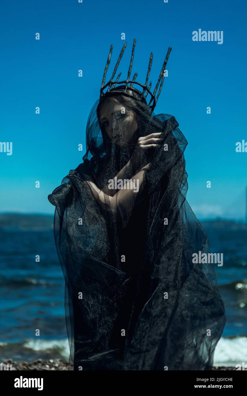 Model wearing black gothic dress and dark crown is posing with a lighthouse Stock Photo