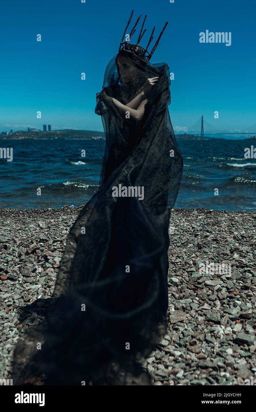 Model wearing black gothic dress and dark crown is posing with a lighthouse Stock Photo