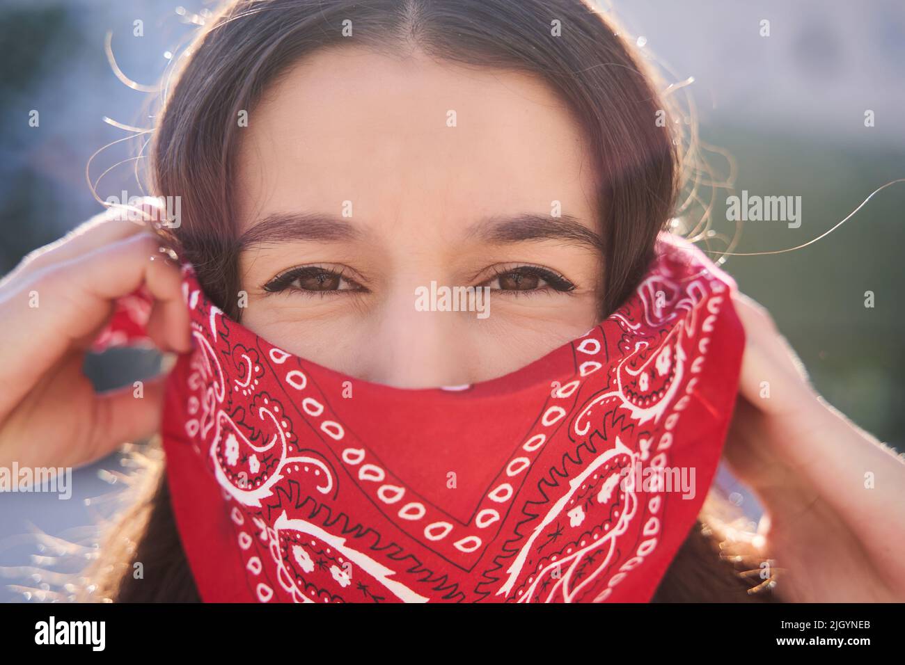 Young brunette woman wearing red bandana covering half of face, looking at camera, activist protest concept. Stock Photo