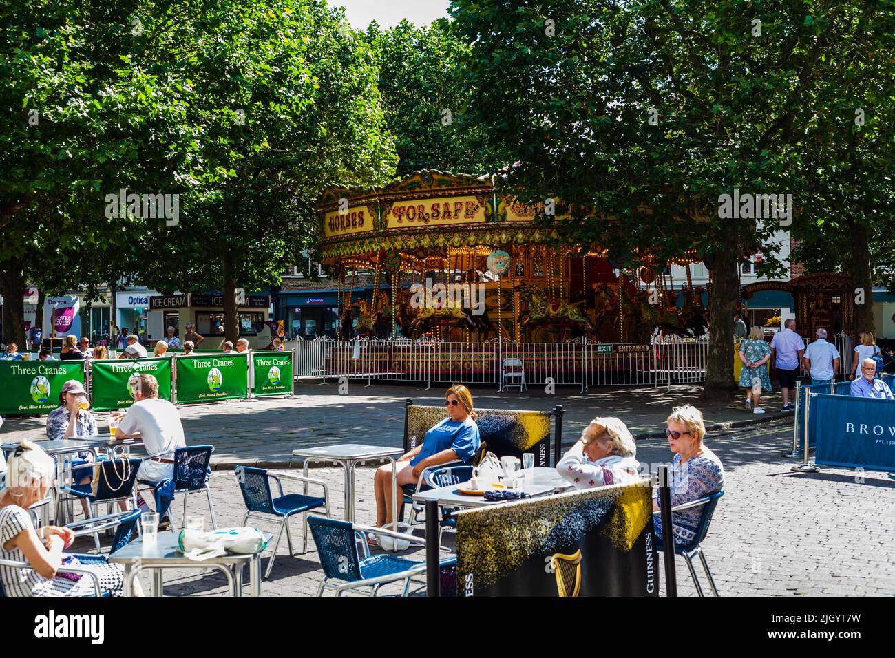 People sat outside a restaurant in the morning sunshine with a fairground roundabout in background in York,North Yorkshire,England,UK Stock Photo