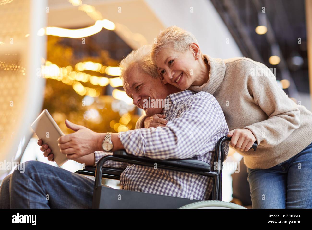 Happy loving attractive mature wife hugging person with disability husband in wheelchair while he watching video on tablet Stock Photo