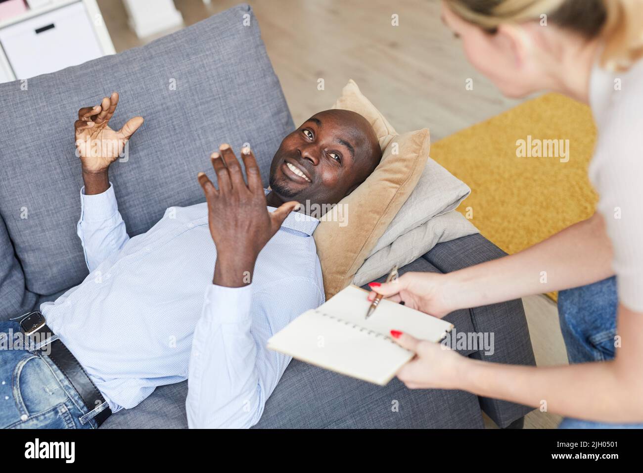 Smiling excited young black man with bald head lying on comfortable sofa and describing his dream cheerfully while talking to therapist at therapy ses Stock Photo