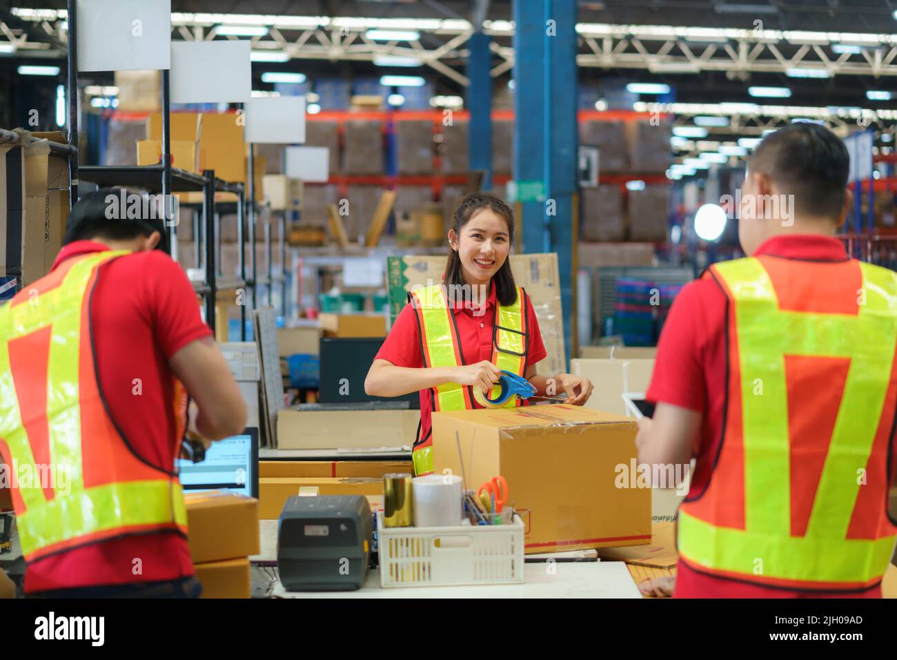 Asian female worker packing cardboard box with tape gun dispenser in warehouse. Thai employee packing goods in large industrial storehouse. Stock Photo