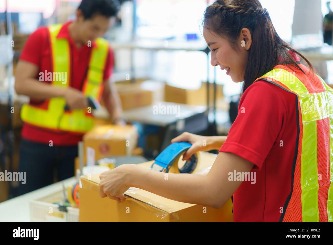 Asian female worker packing cardboard box with tape gun dispenser in warehouse. Thai employee packing goods in large industrial storehouse. Stock Photo