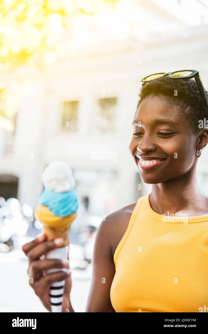 portrait of an african woman looking the colorful ice-cream on her hand. She wears summer attire and smiles Stock Photo