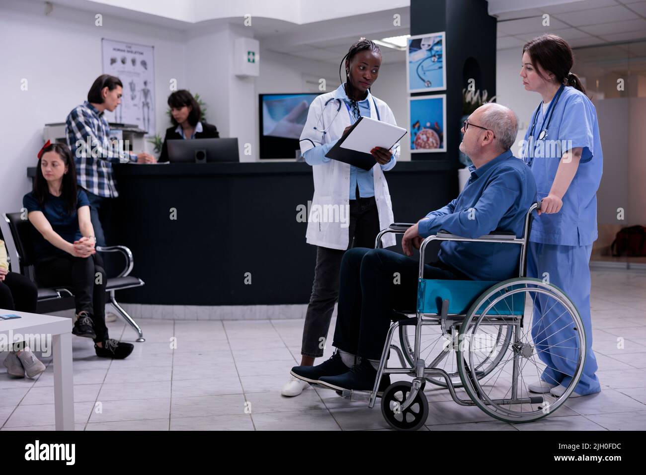 Older man living with disability being helped by caring nurse talking with doctor holding clipboard with patient history. Senior citizen using wheelchair in clinic reception with diverse people. Stock Photo