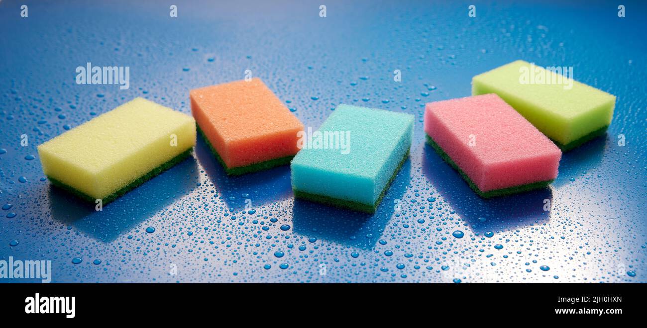 Collection of multicolored wash sponges placed on wet blue background covered with drops of water in light room during household routine Stock Photo