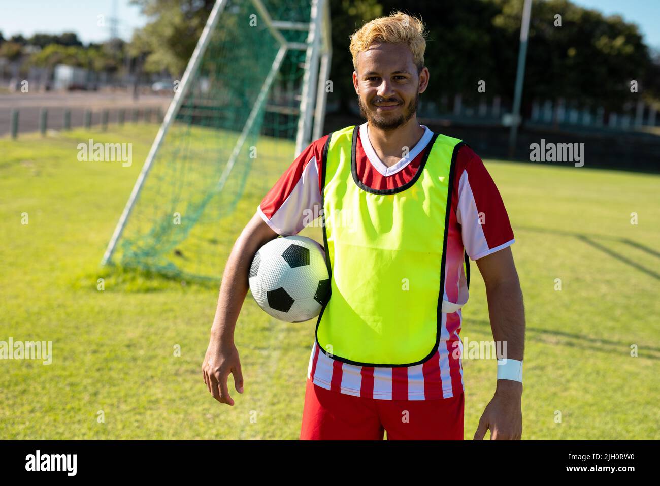 Portrait of smiling caucasian young player in red jersey with soccer ball standing in playground Stock Photo