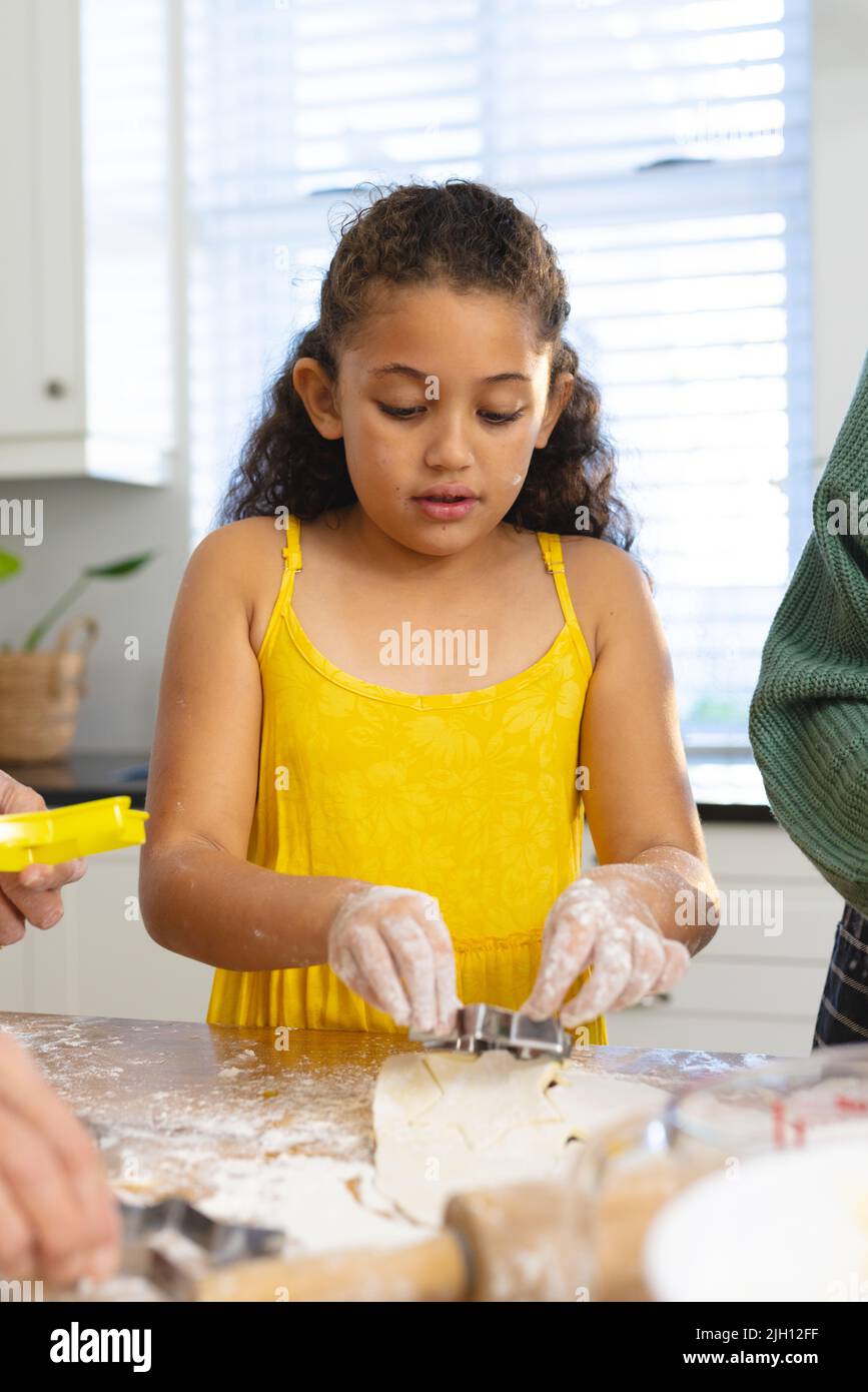Cute multiracial girl with messy hands cutting dough with pastry cutter on table in kitchen Stock Photo