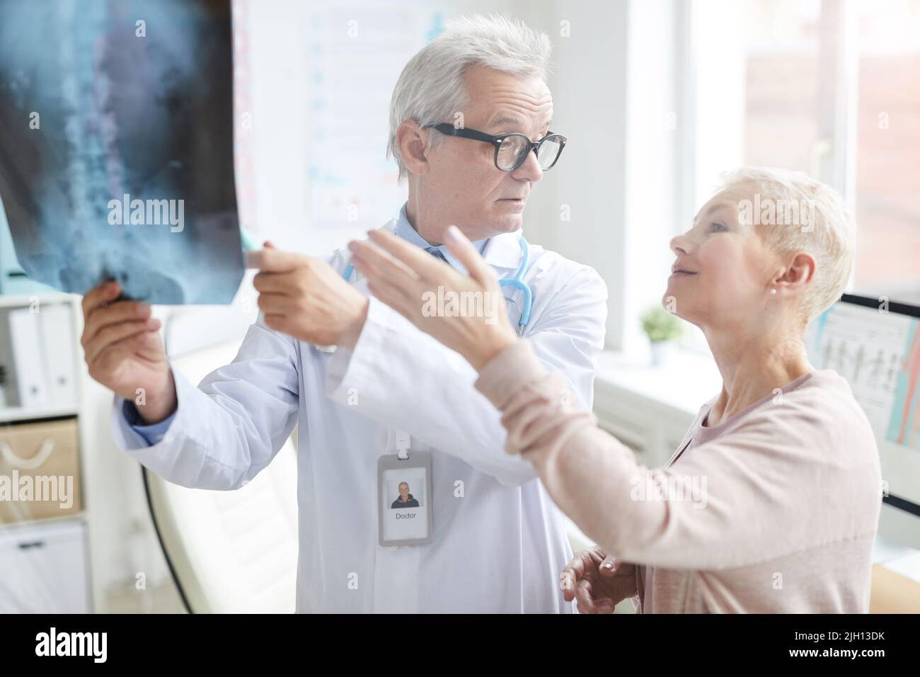 Attractive blond-haired mature lady pointing at core x-ray image while discussing her pain with doctor Stock Photo