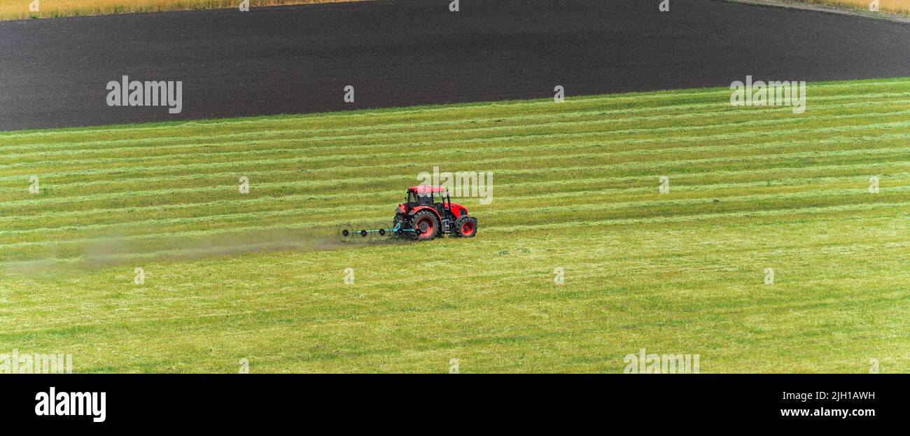 Red tractor mowing on green cultivated agriculture green field, panoramic image. Stock Photo