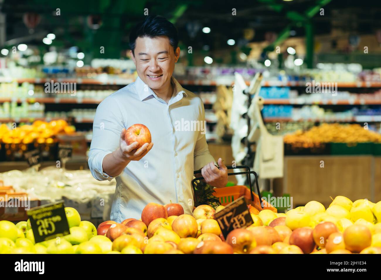 young asian man chooses and picks in eco bag apple fruit or vegetables in the supermarket. male customer standing a grocery store near the counter buys and throws in a reusable package in market Stock Photo