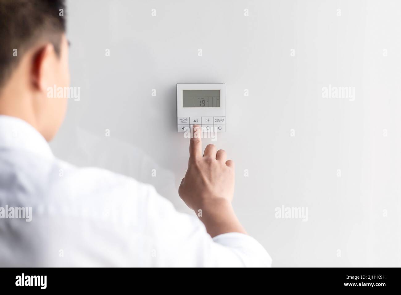 Close-up photo. The hand of a young man in a white shirt turns on the control buttons of the air conditioner hanging on the wall. Standing on the left Stock Photo
