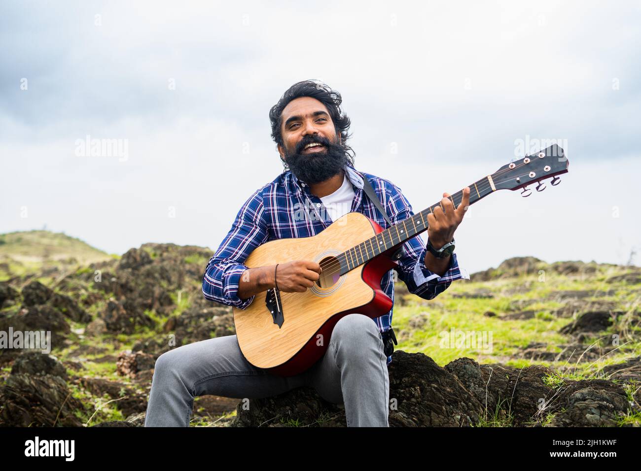 Young beard man playing guitar by singing song while sitting on top of hill - concept of hobbies, leisure activity and entertainment Stock Photo