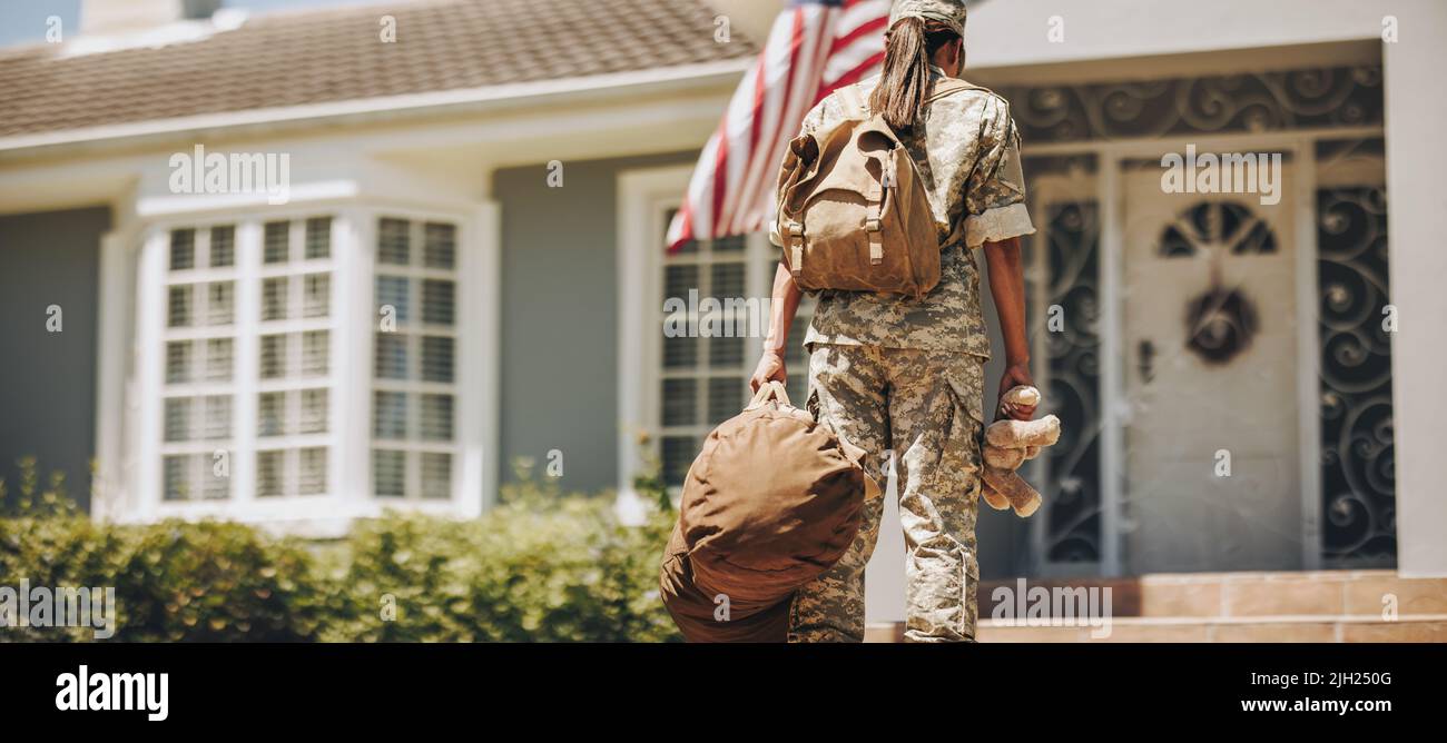 Back view of a military mom holding a teddybear while standing outside her house with her luggage. Courageous female soldier coming back home after se Stock Photo