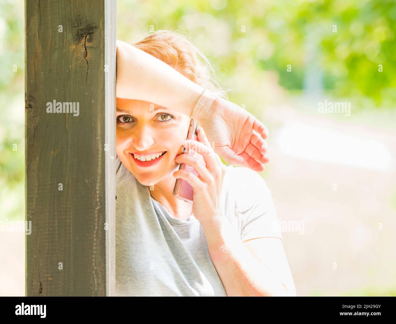 Eyeshot blonde hair woman 30s is talking on smartphone inclined on wooden post column happy smiling looking at camera high-hey highkey Stock Photo
