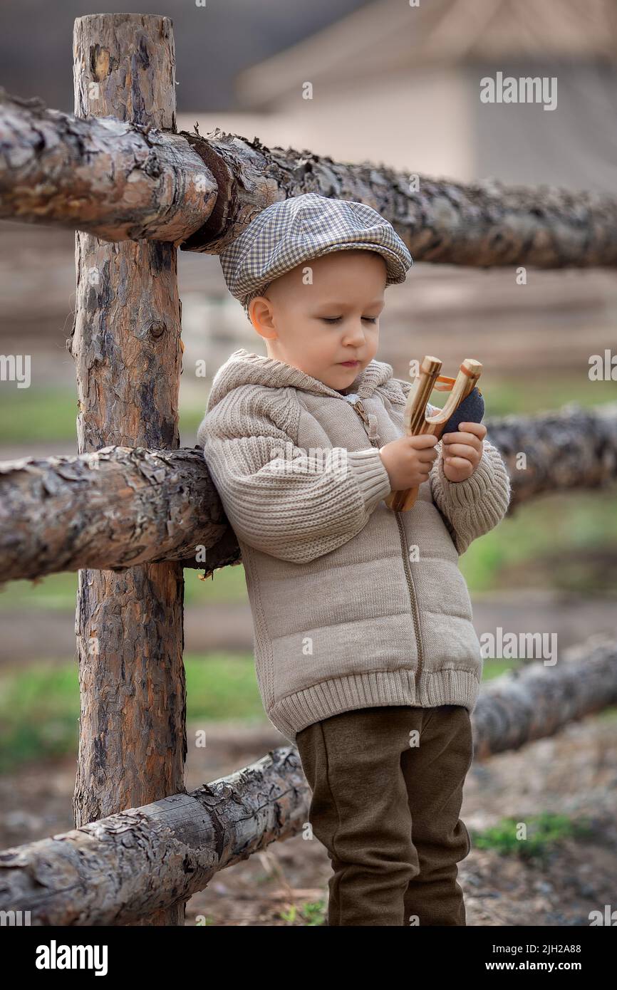 Bully boy kid with a slingshot aims at someone near a fence in the village outdoors. Rustic barefoot child boy with a hat shoots a slingshot. Stock Photo