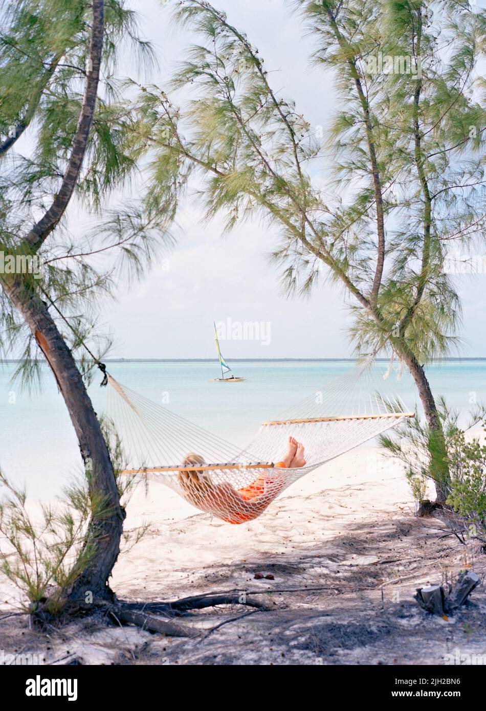 A guest relaxes in a hammock on Tiamo Beach as a catamaran sails by. South Andros Island, Bahamas Stock Photo