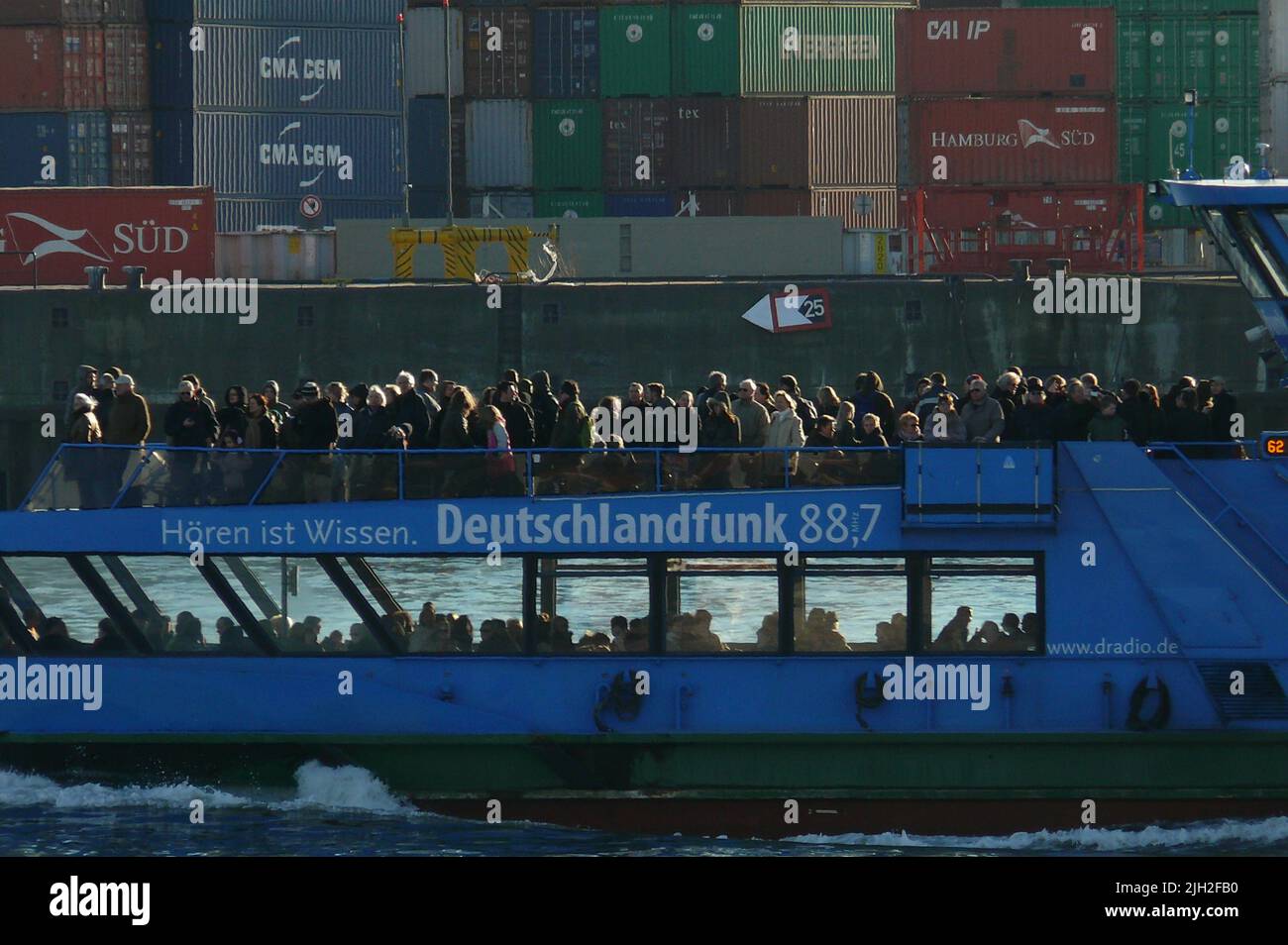 Eine Hafenfähre der Linie 62 fährt in Hamburg an einem Containerfrachter vorbei.Das Deck der Hafenfähre ist  voller Fahrgäste und Touristen. Die Fähre der Linie 62 wird gerne als preiswerte Hafenrundfahrt genutzt . An Deck des Containerfrachters stapeln sich Container in vielen Farben von verschiedenen Reedereien. Stock Photo