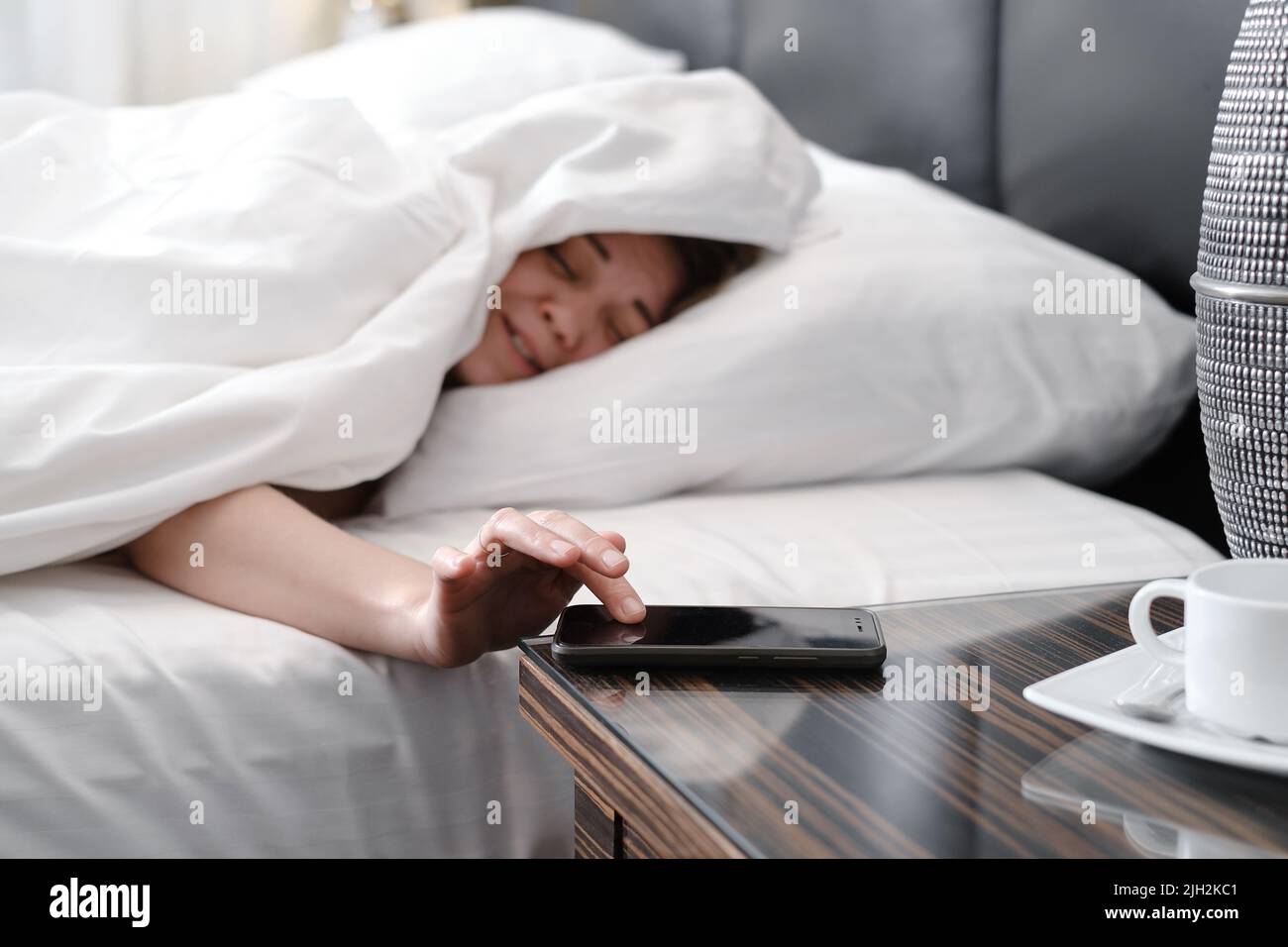 Sleepy Woman covered with blanket in the morning in bed turning off alarm clock On Cell Phone. Hard to wake up early from bed Stock Photo