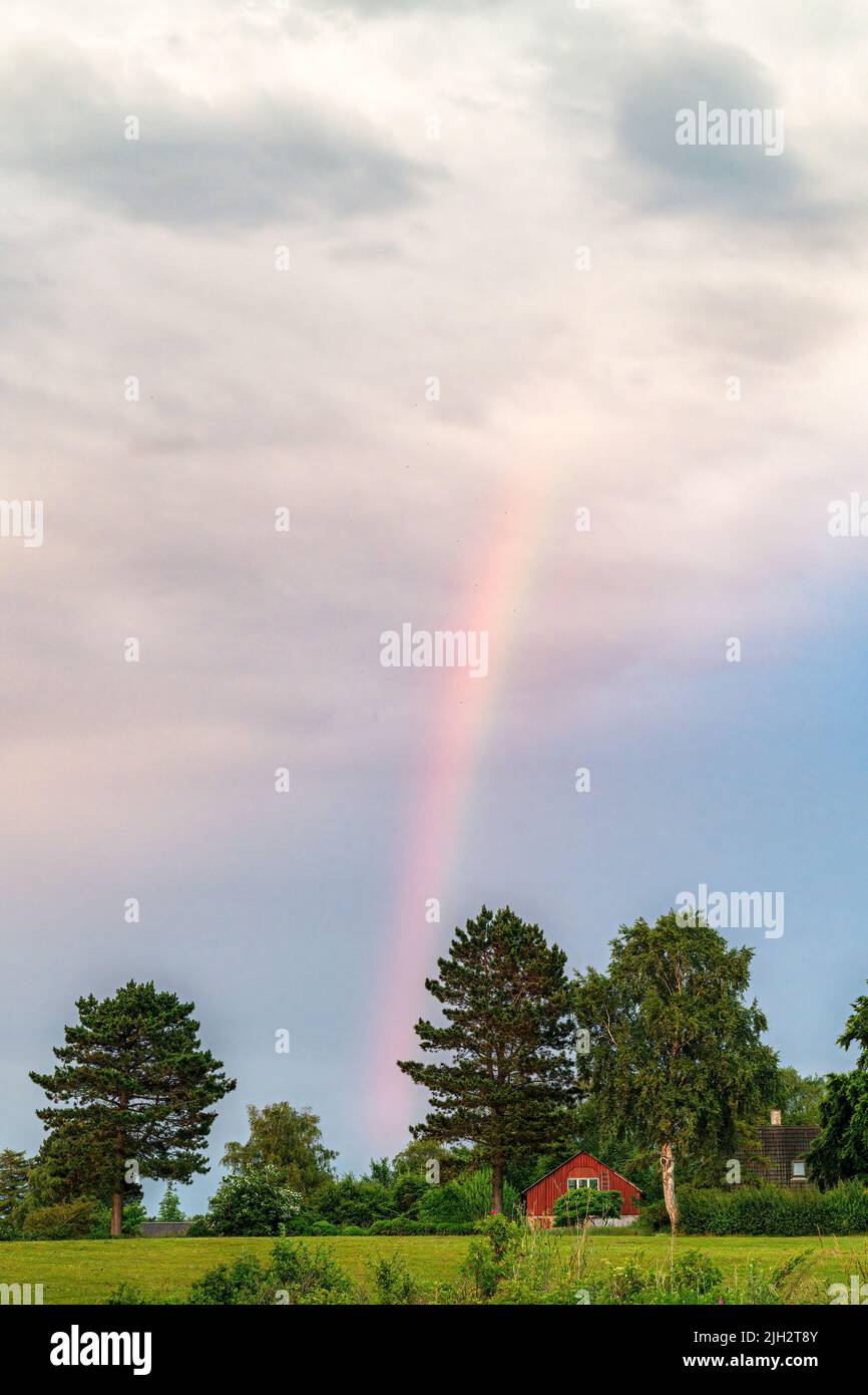A rainbow dominates the sky over a red wooden country house. Stock Photo