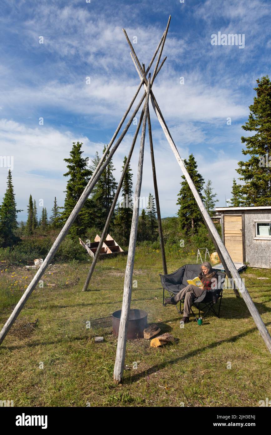 Woman sitting at firepit reading beneath a teepee structure in summer, near the northern Indigenous community of Deline, Northwest Territories, Canada Stock Photo