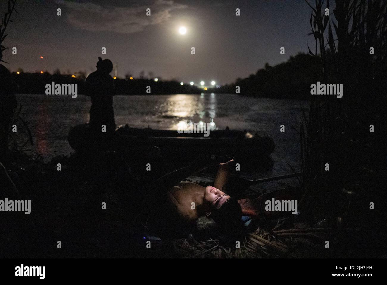 The Buck Moon rises in the background as a smuggler, illuminated by his phone, takes rest on a sandbar between smuggling migrants across the Rio Bravo del Norte, also known as the Rio Grande river, into the United States from Ciudad Miguel Aleman, Mexico July 13, 2022. REUTERS/Adrees Latif     TPX IMAGES OF THE DAY Stock Photo
