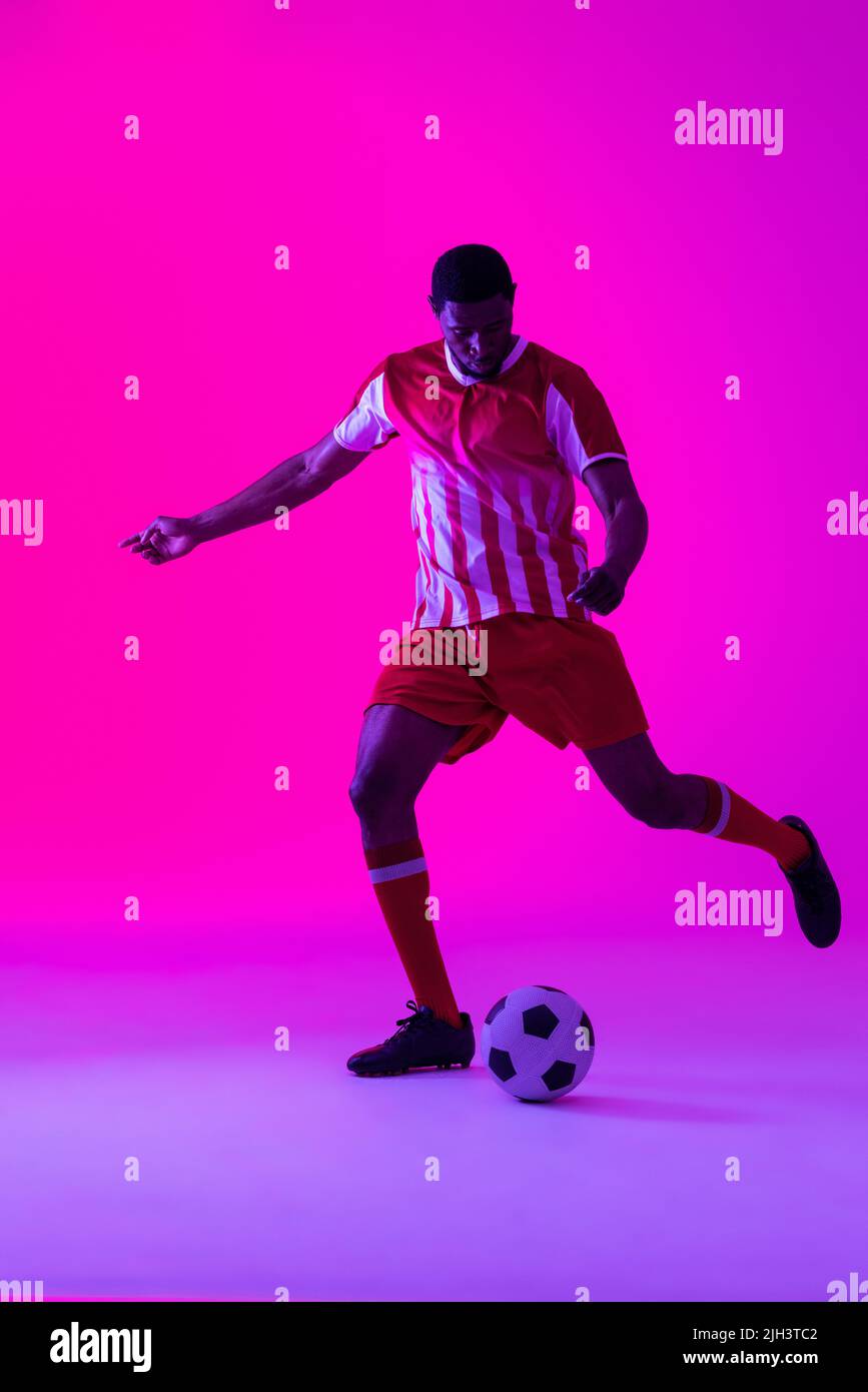 African american male soccer player kicking football over neon pink