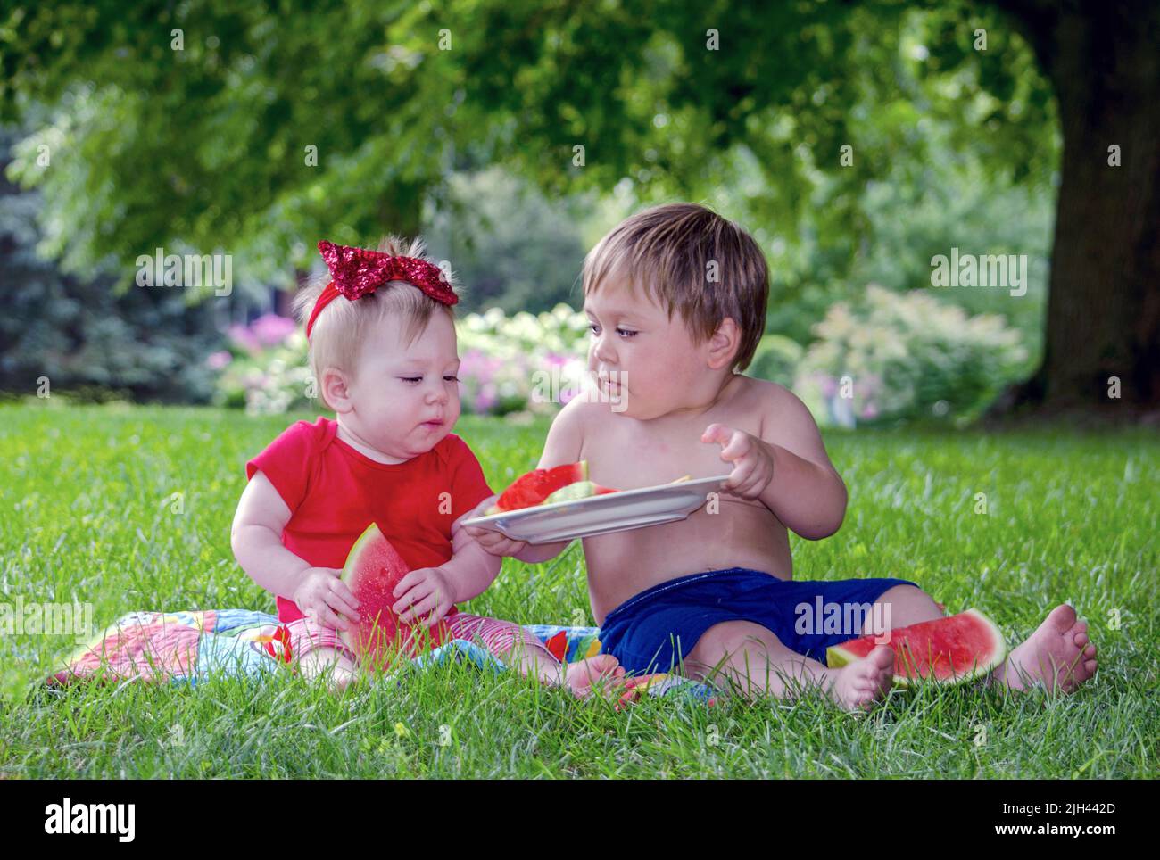 Young toddler boy shares watermelon with his baby sister as they sit outside on a summers day Stock Photo