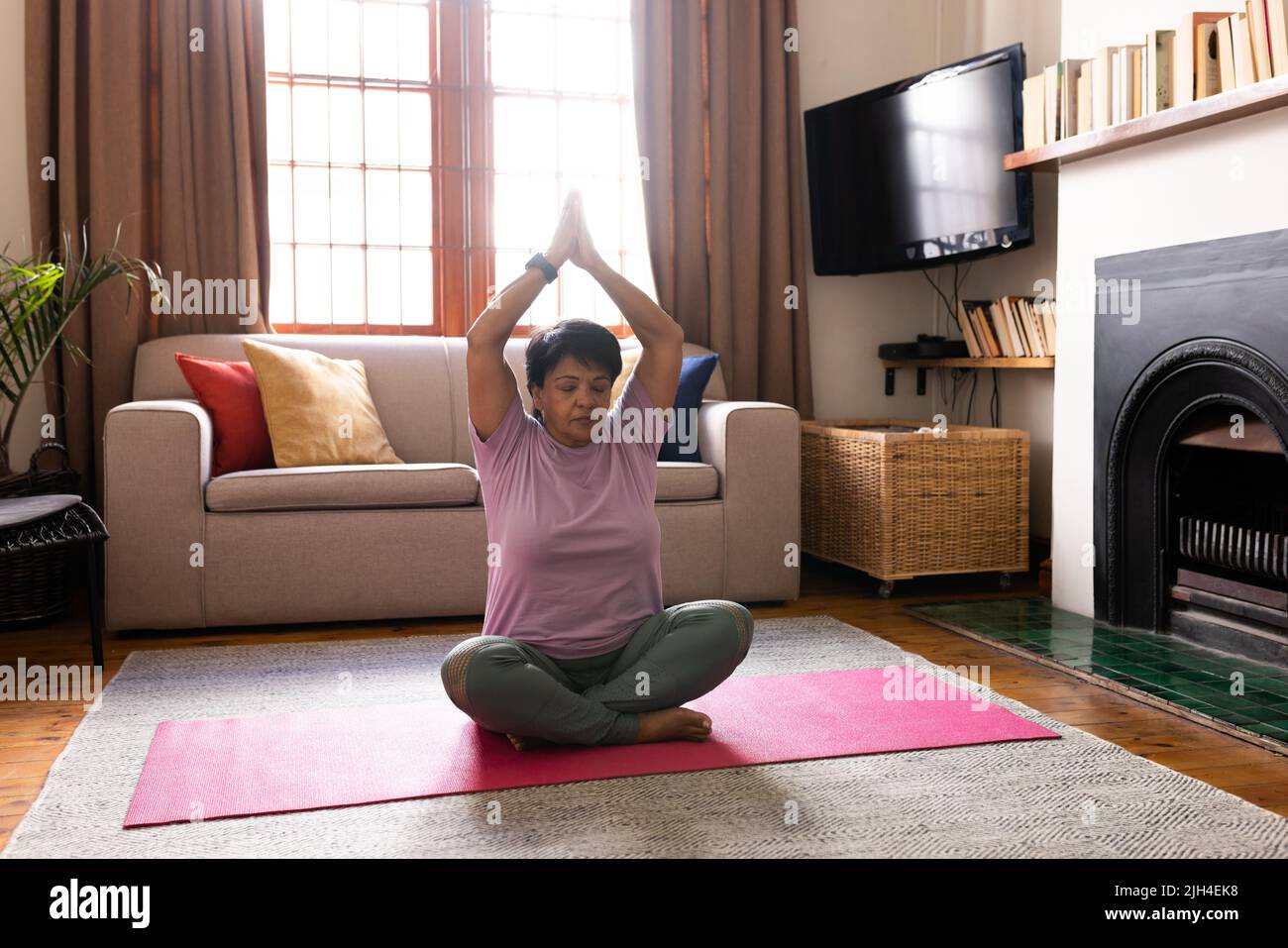 Biracial mature woman with arms raised and eyes closed meditating while sitting on mat at home Stock Photo