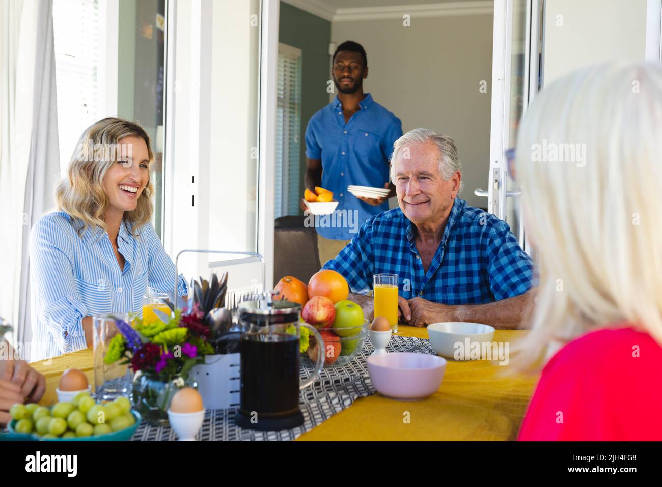 Multiracial man with plates and food standing at doorway while family talking and having breakfast Stock Photo
