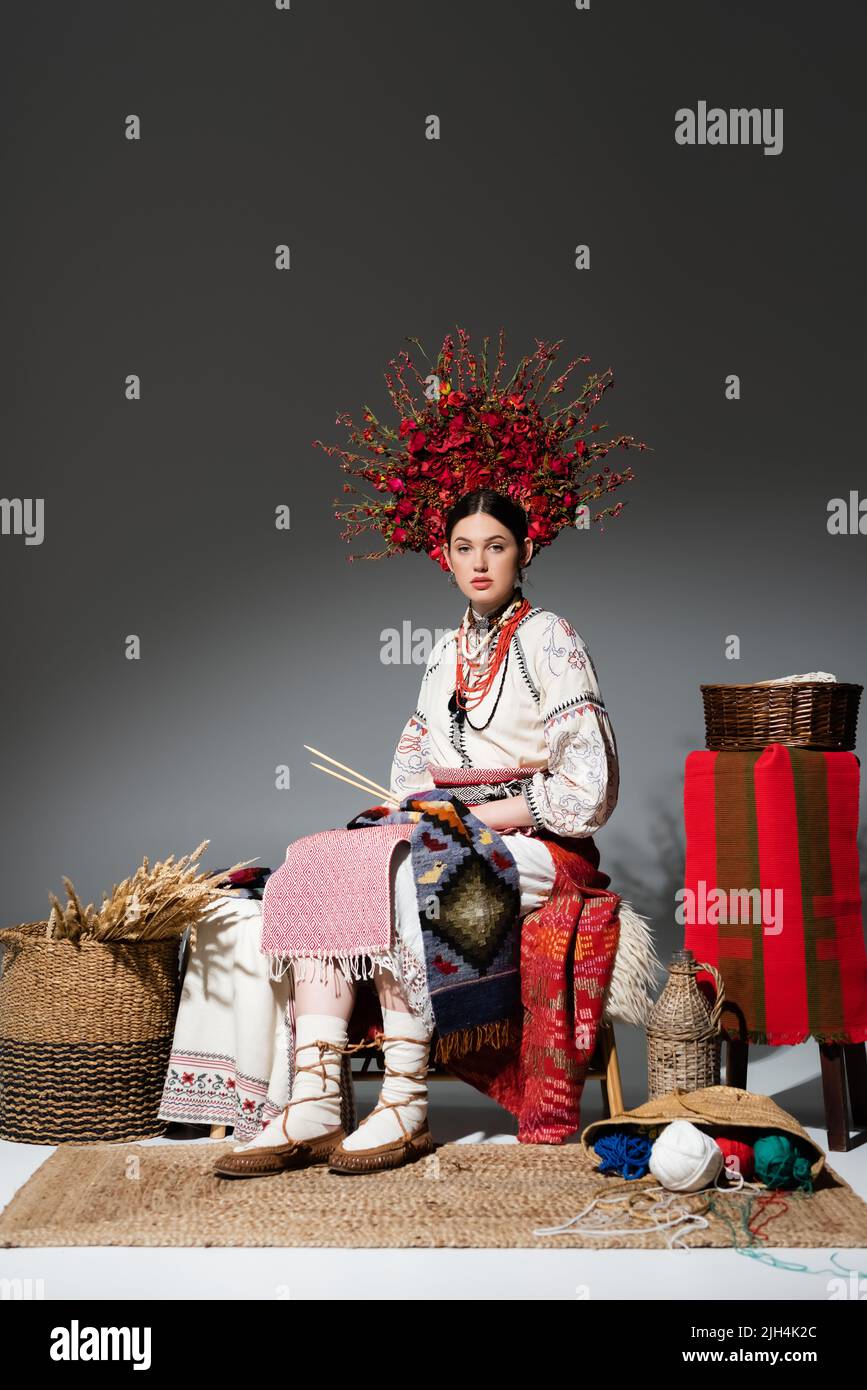 full length of brunette ukrainian woman in traditional clothes and wreath with flowers holding knitting needles on dark grey Stock Photo