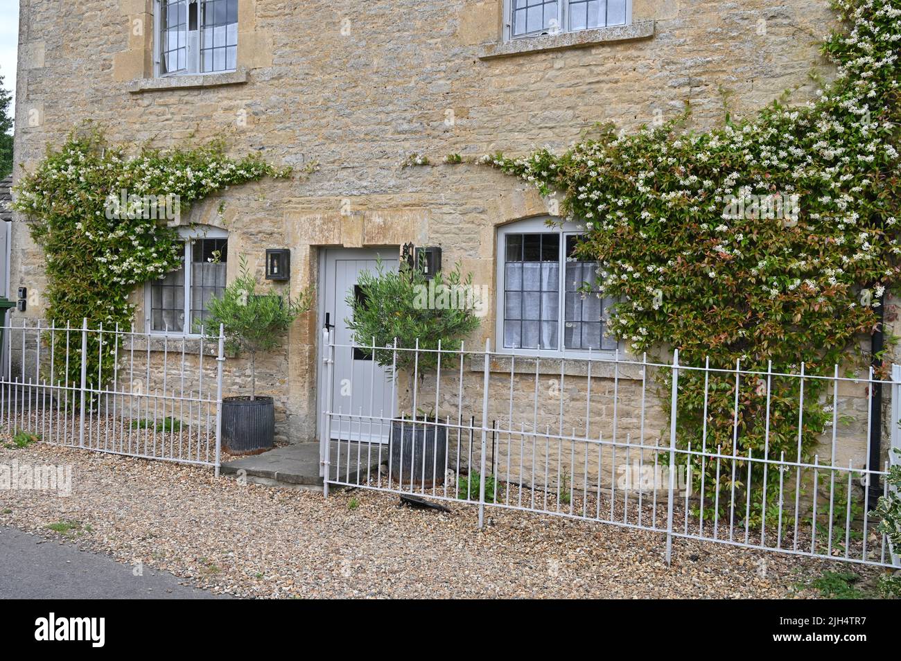 The front of a house in the Gloucestershire vilalge of Upper Slaughter with jasmine growing up the wall. The gaden is enclosed by railings. Stock Photo