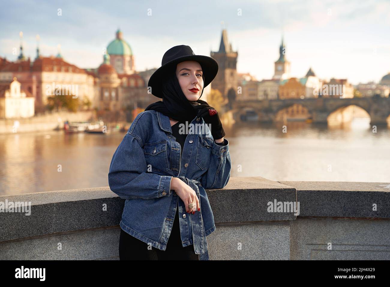 Stylish beautiful young woman earing black hat in Prague with Charles Bridge on background. Elegant retro lady fine art portrait. Stock Photo