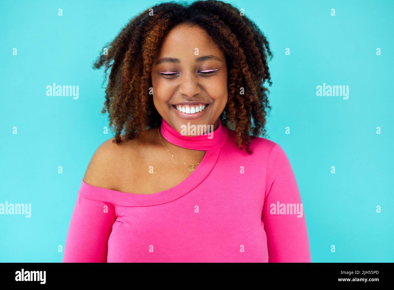Dreamy young beautiful African American woman with bright eyelines wearing pink bodysuit over blue wall keeps her eyes closed, looks with happy expres Stock Photo