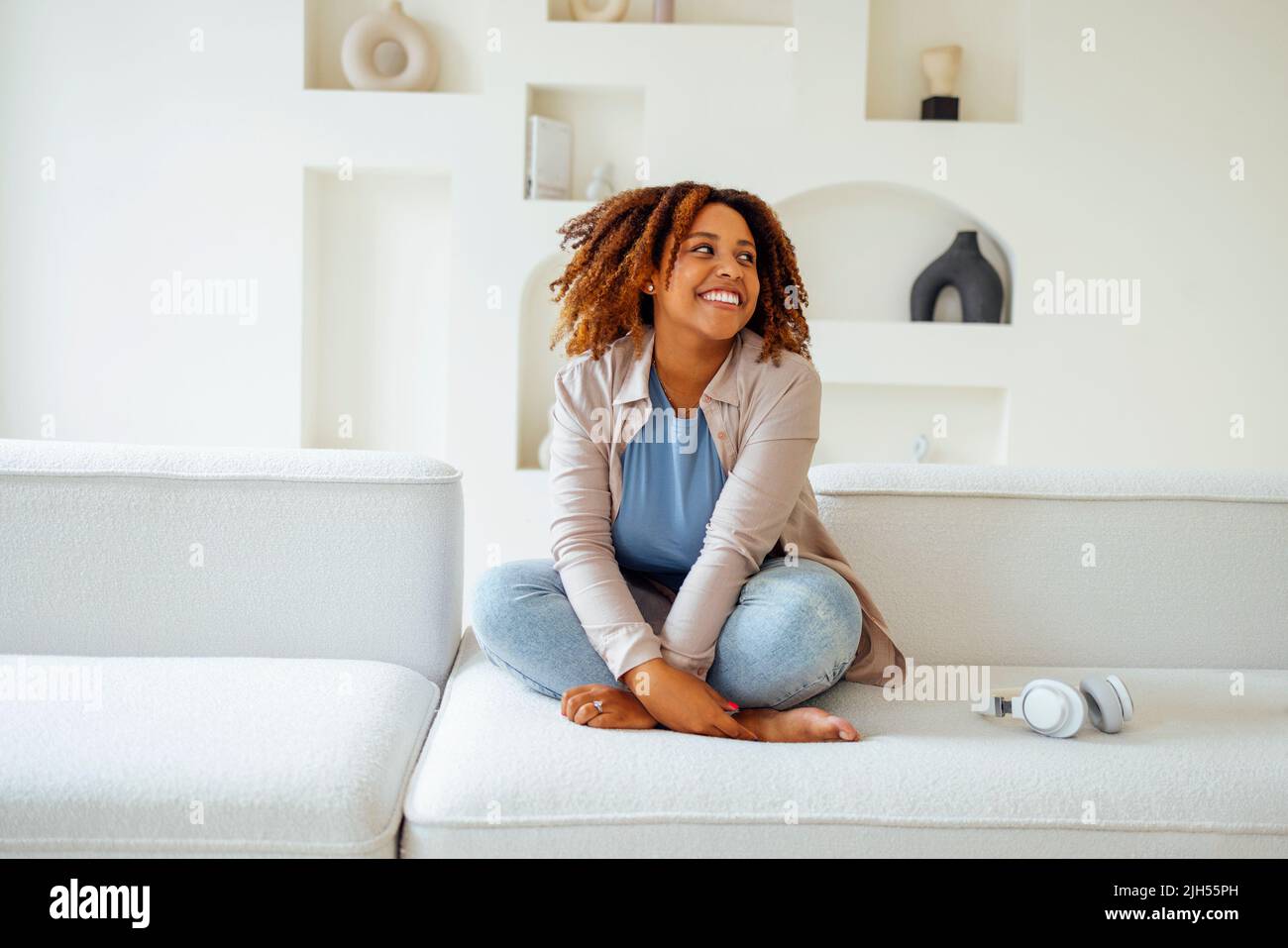 African american millennial girl, contented and happy, sit on comfortable sofa in cozy modern light living room. Enjoy her live Stock Photo