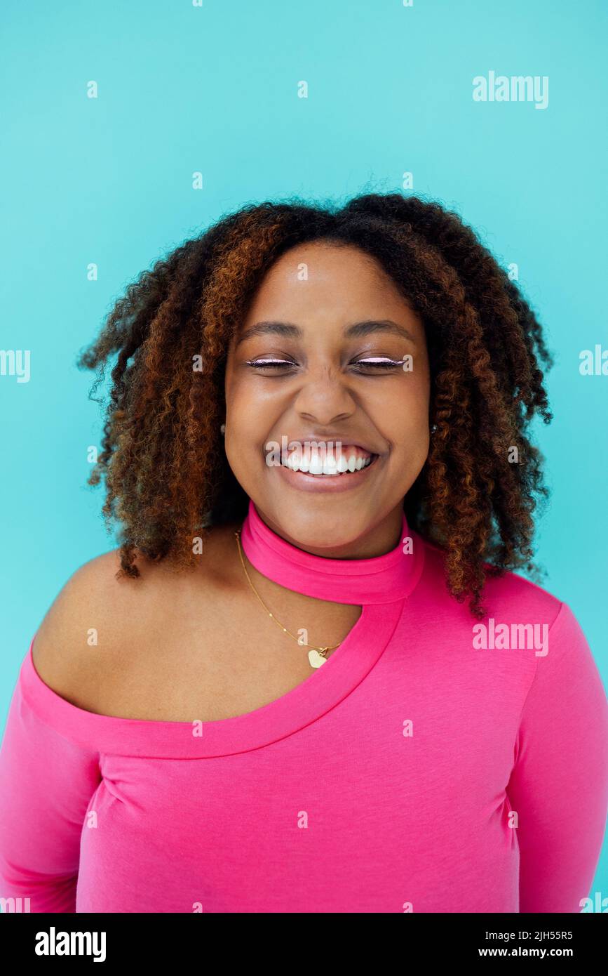 Dreamy young beautiful African American woman with bright eyelines wearing pink bodysuit over blue wall keeps her eyes closed, looks with happy expres Stock Photo