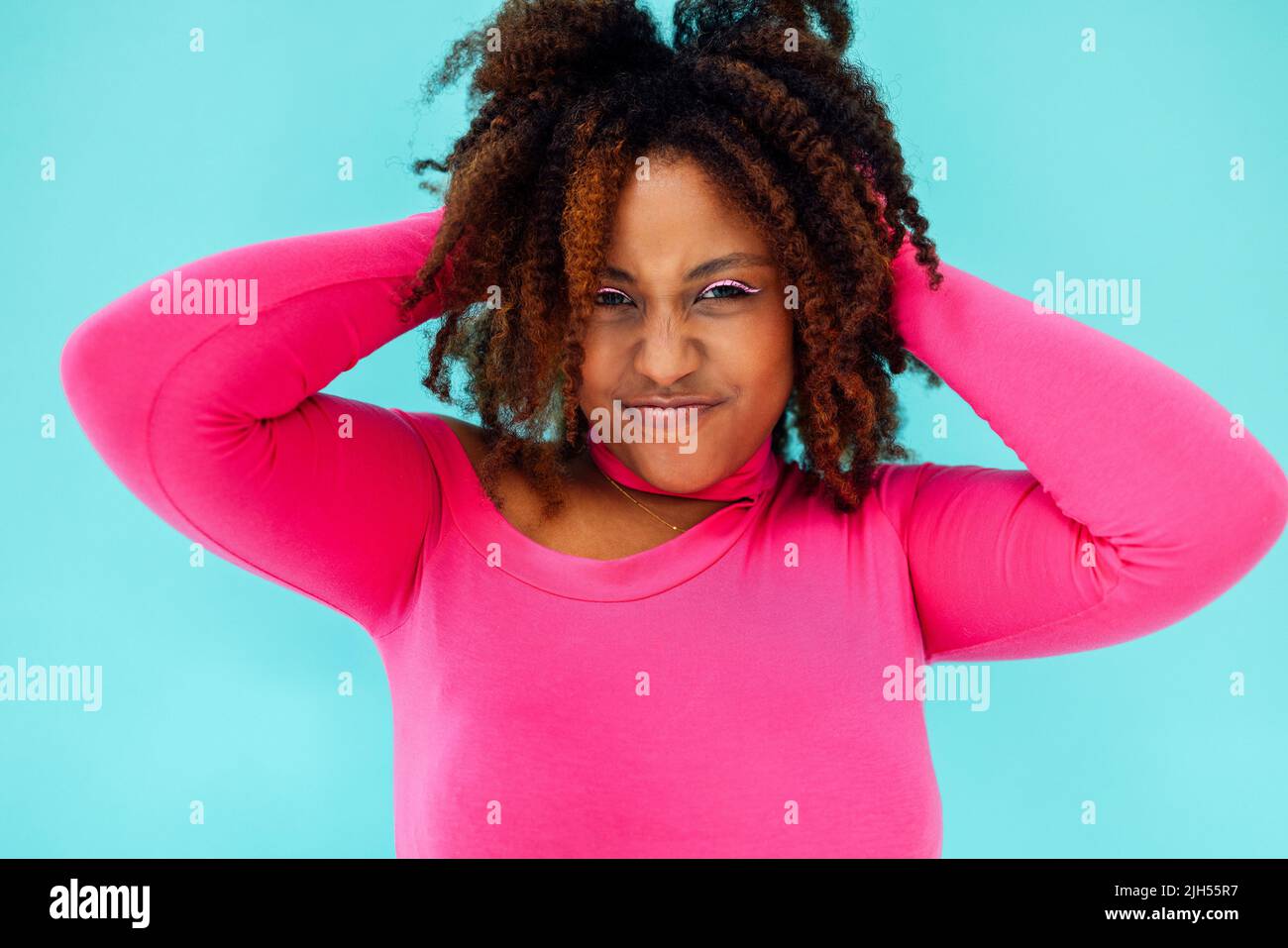 Dreamy young beautiful African American woman with bright eyelines wearing pink bodysuit over blue wall keeps hands pressed together under chin, looks Stock Photo