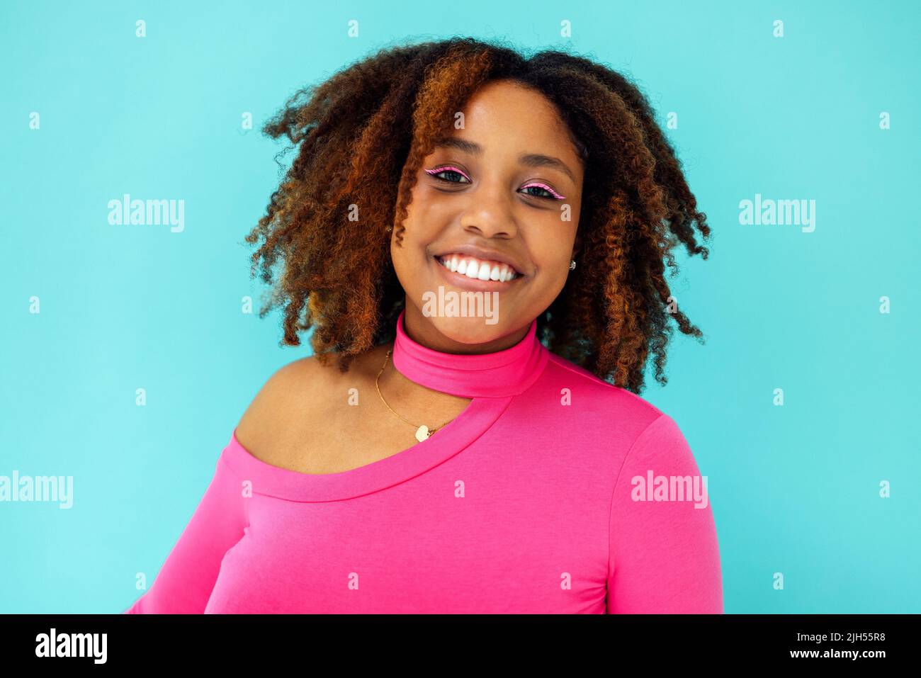 Dreamy young beautiful African American woman with bright eyelines wearing pink bodysuit over blue wall, looks with happy expression, has toothy smile Stock Photo
