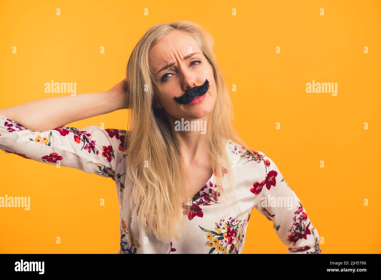 Funny long-haired blond caucasian woman in her mid 20s with fake black moustache on her face. Confused facial expression. Medium closeup studio shot over orange background. High quality photo Stock Photo