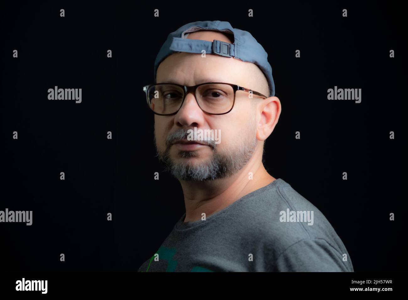 Portrait of a bald bearded man wearing cap and glasses looking at the camera against a black background. Salvador, Bahia, Brazil. Stock Photo