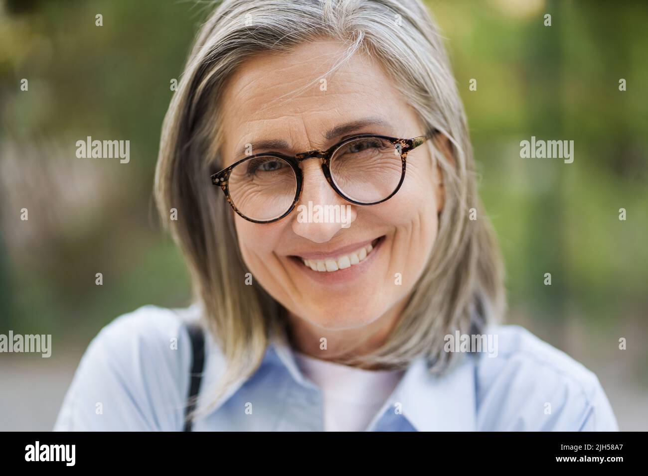 Close up portrait of charming grey hair mature woman in glasses standing outdoor enjoying free time vacation traveling around world at retirement. Mature woman with perfect skin wearing blue shirt. Stock Photo