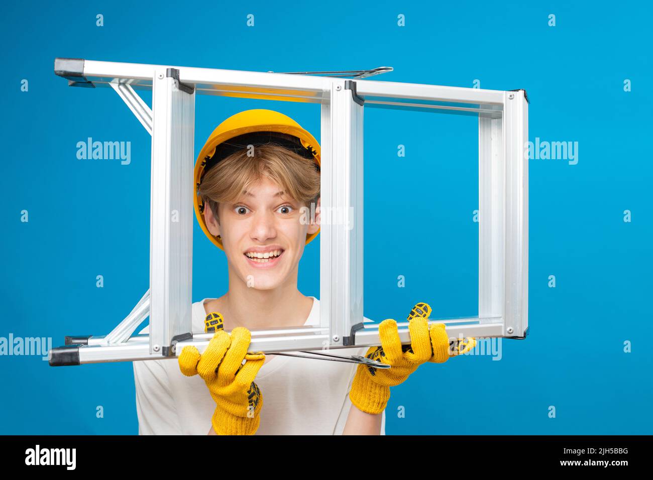 Happy teen in white t-shirt and gloves with yellow helmet in head, holding ladder in hands standing in studio on blue background. Stock Photo