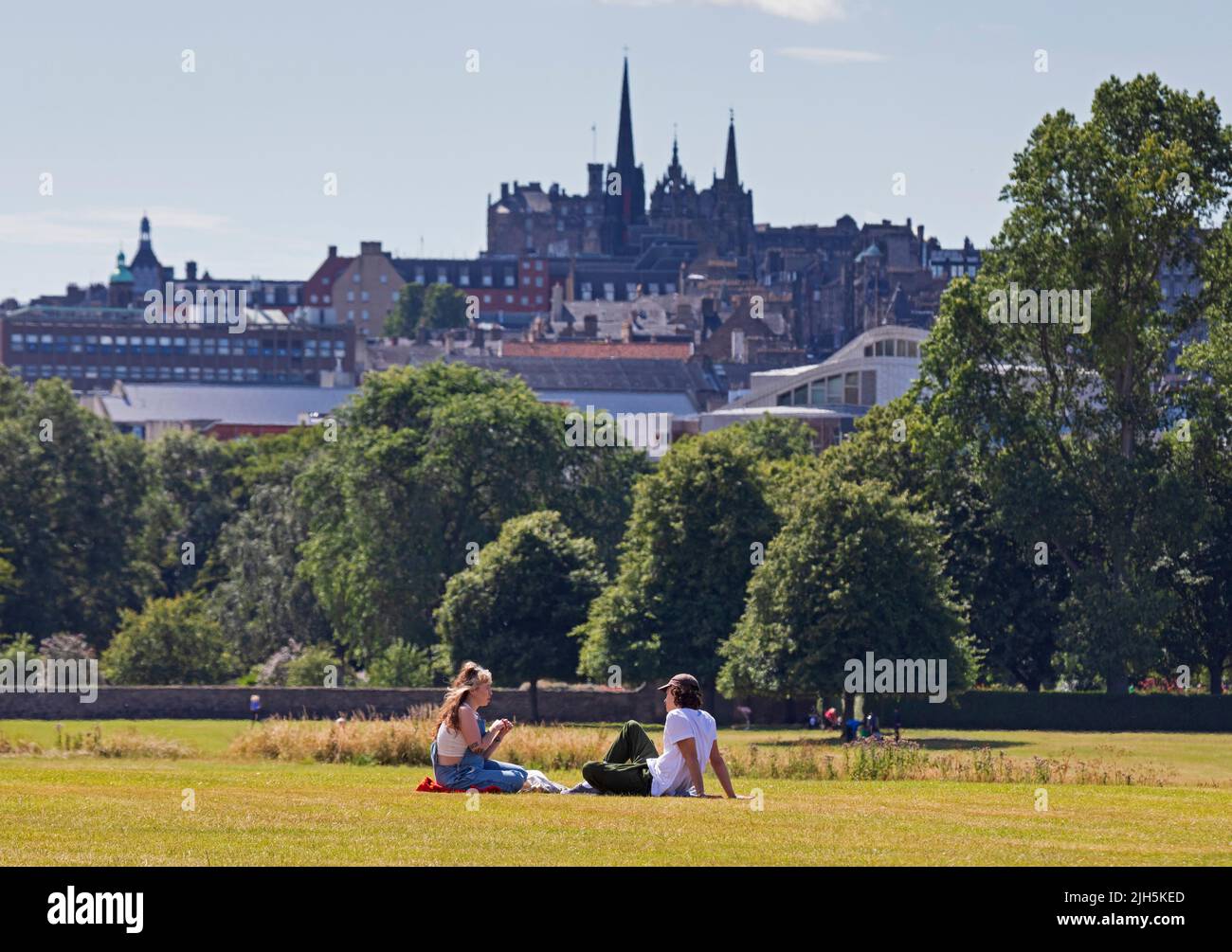 Holyrood Park, Edinburgh, Scotland, UK. 15th July 2022. People enjoy the summer in the city with temperature of 21 degrees centigrade in the scenic surroundings of historic Holyrood Park in the city centre below Arthur's Seat. Pictured: A young couple relax by sitting on the grass chatting with the architecture of the city centre in the background. Credit: Scottishcreative/alamy live news. Stock Photo
