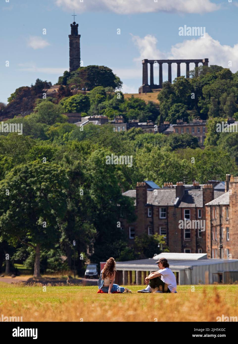 Holyrood Park, Edinburgh, Scotland, UK. 15th July 2022. People enjoy the summer in the city with temperature of 21 degrees centigrade in the scenic surroundings of historic Holyrood Park in the city centre below Arthur's Seat. Pictured: A young couple relax by sitting on the grass chatting with the architecture of the Calton Hill in the background. Credit: Scottishcreative/alamy live news. Stock Photo