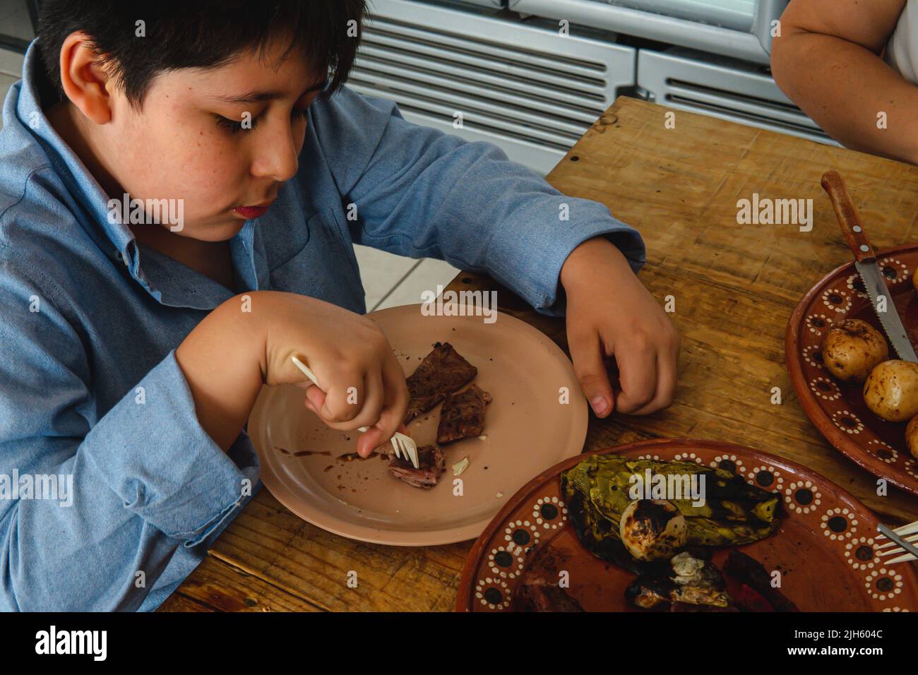 Top view of an elementary school boy with various dishes on the table eating a cut of meat. High quality photo. Wooden table. A plate of clay with food. Children wearing a blue shirts.  Stock Photo