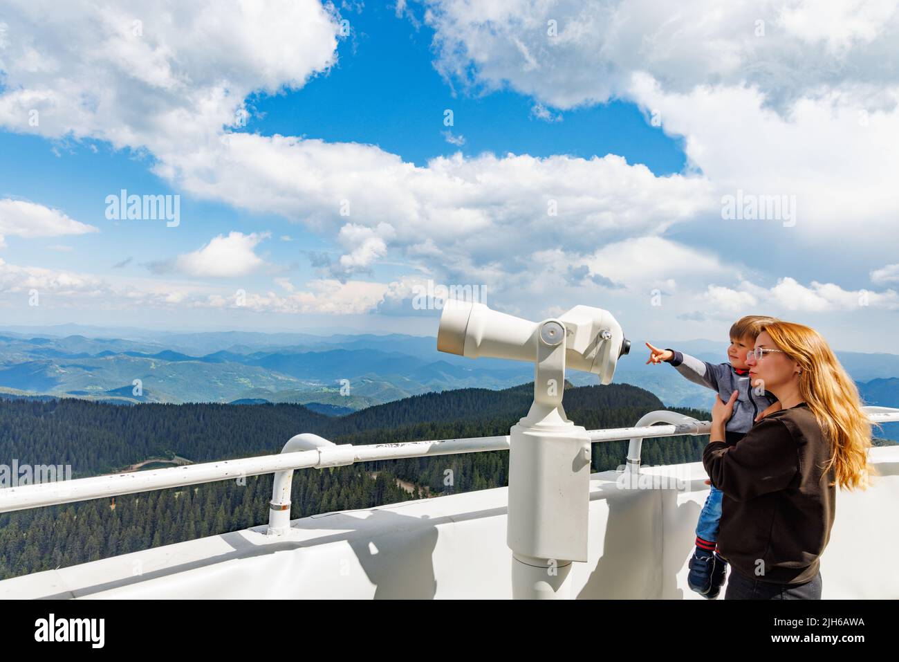 A caring mother shows her curious baby son the landscapes in the forested valley of the Rhodope Mountains and the cloudy sky through a telescope, on a Stock Photo