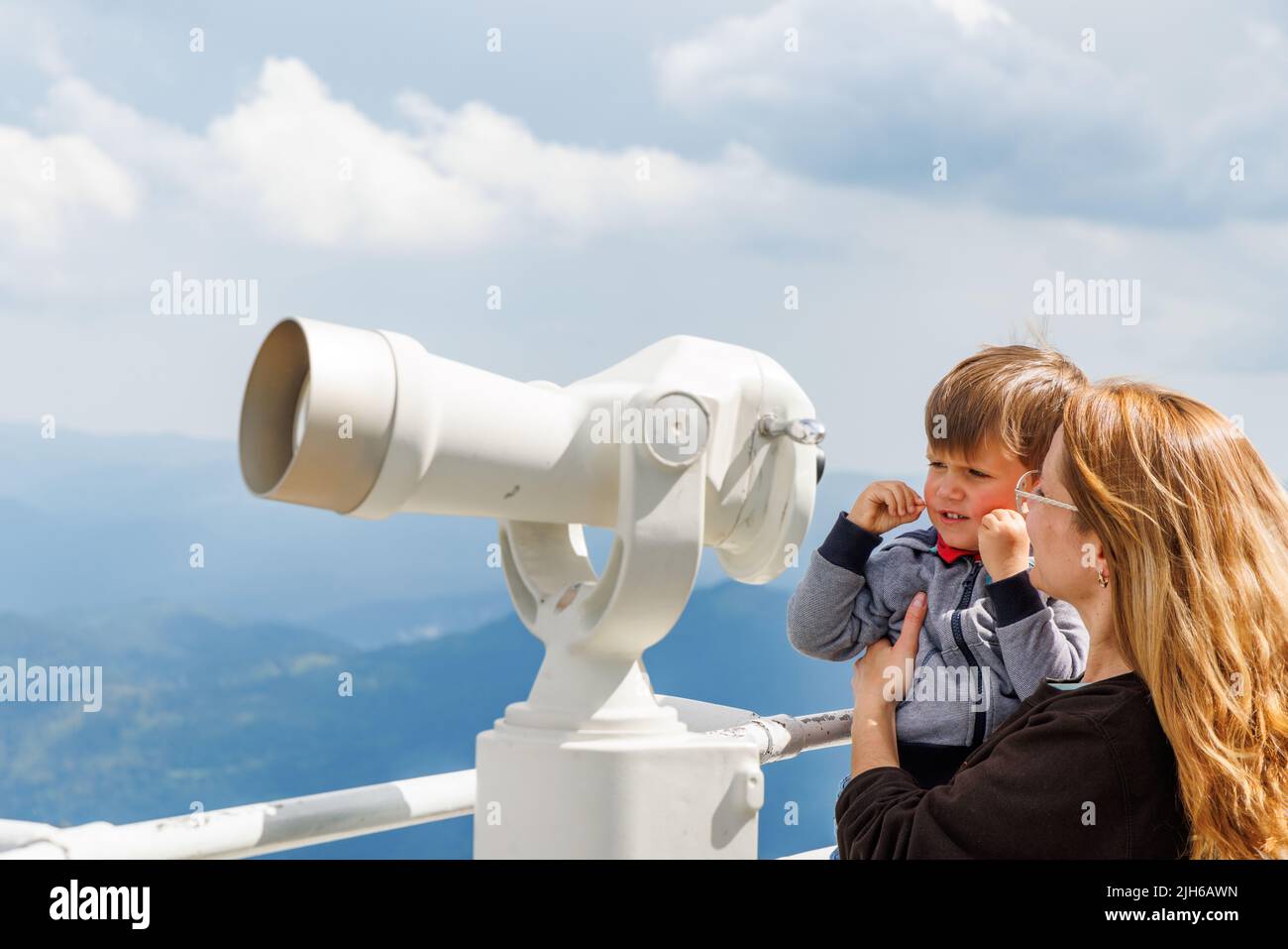 A caring mother shows her curious baby son the landscapes in the forested valley of the Rhodope Mountains and the cloudy sky through a telescope, on a Stock Photo