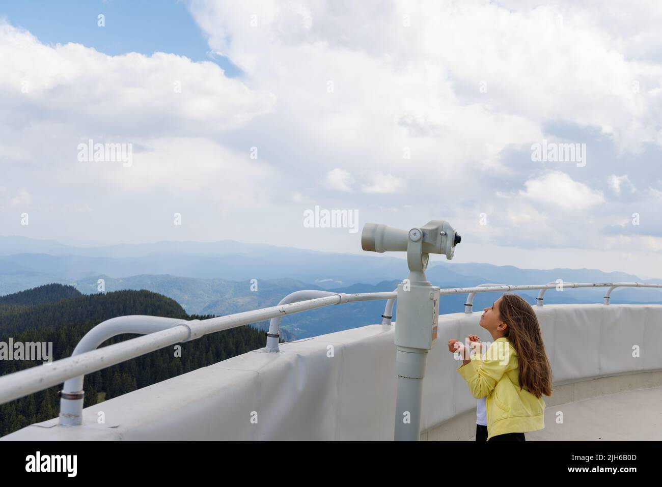 Curious little girl looking at landscapes of mountain forest valley of Rhodope Mountains and bright blue cloudy sky through modern metal telescope, on Stock Photo