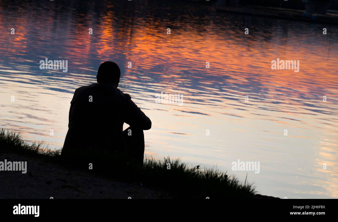 An unidentified lonely man sitting at the evening river bank and anjoying sunset Stock Photo